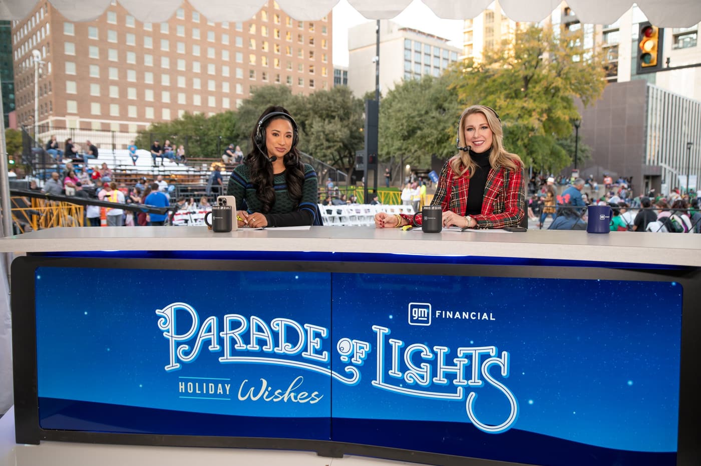 Two women wearing headsets sit behind a desk with a sign that reads 'Parade of Lights Holiday Wishes.' Behind them, a crowd watches from bleachers in front of downtown buildings.