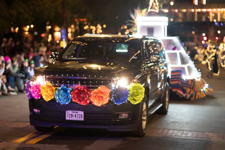 A black vehicle decorated with illuminated rainbow-colored roses along the front grill, driving at night during a parade with spectators on the sidewalk and blurred festive lights in the background.