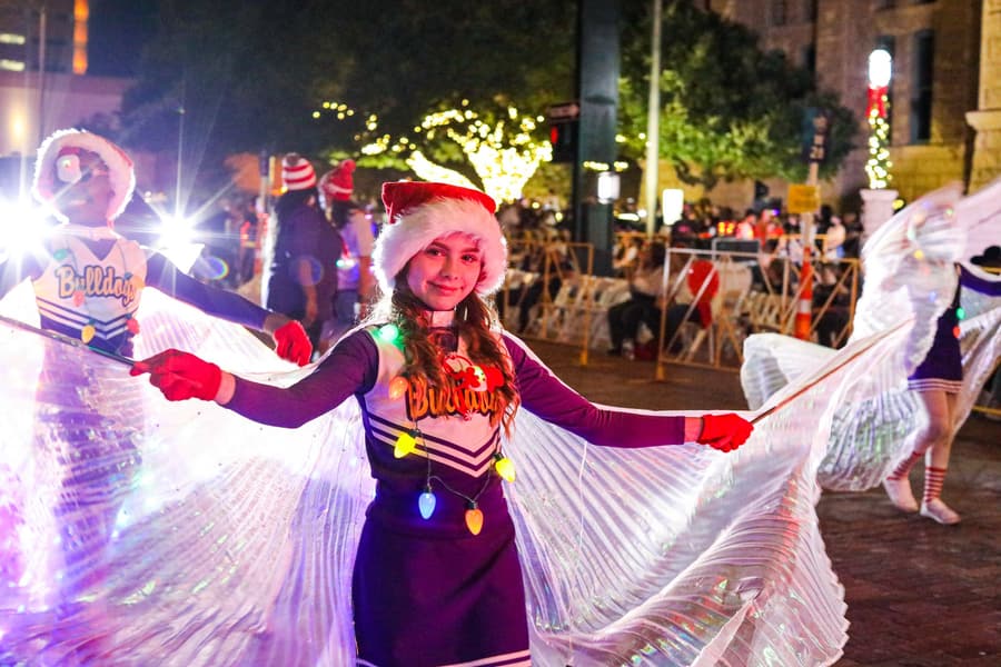 Young woman wearing a Santa hat and a festive costume, holding a shiny, translucent curtain or fabric decorated with colorful Christmas lights at night, with other people in holiday costumes and Christmas lights in the background.