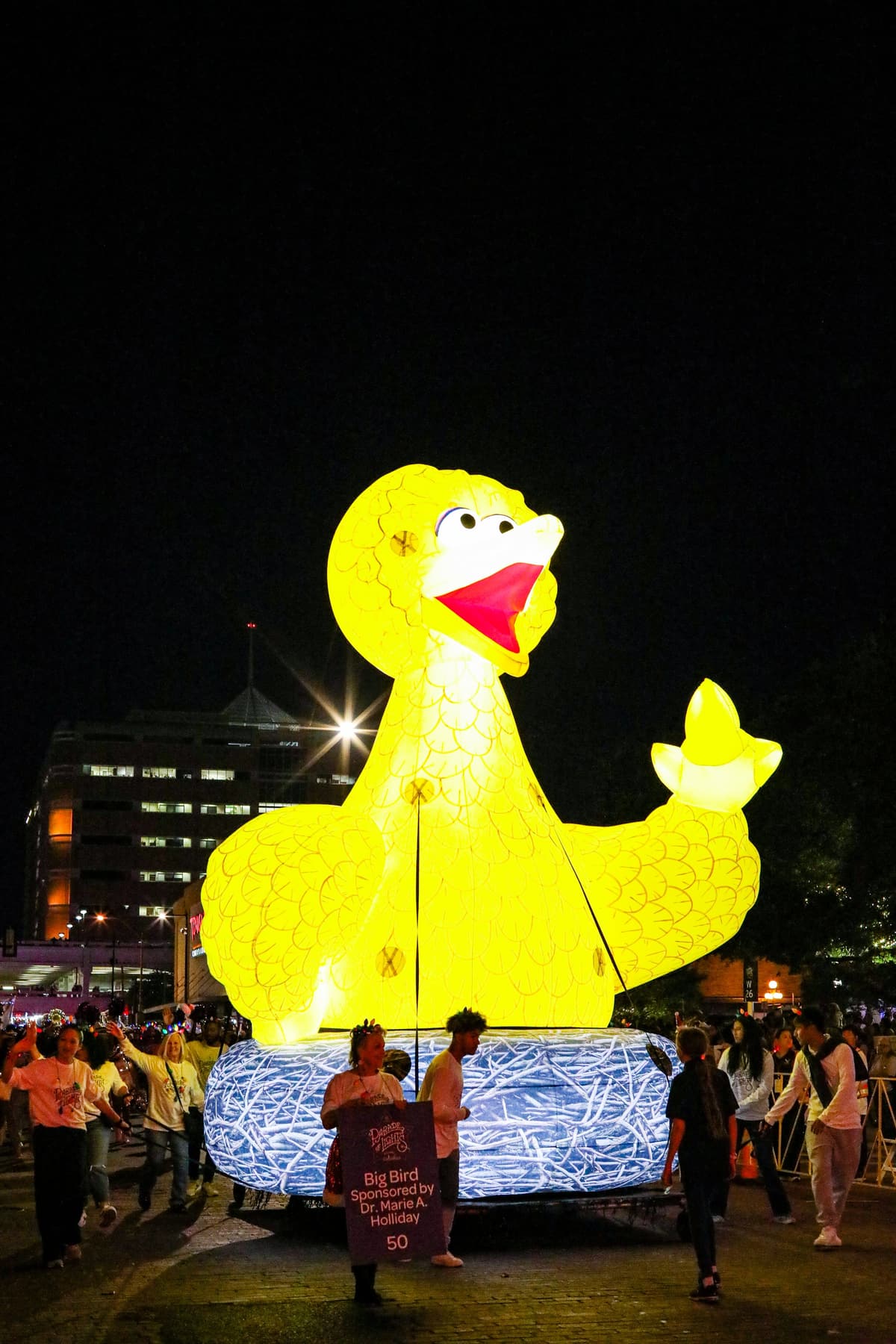 Large illuminated yellow Big Bird lantern with a smiling face, surrounded by people at night in an outdoor event.