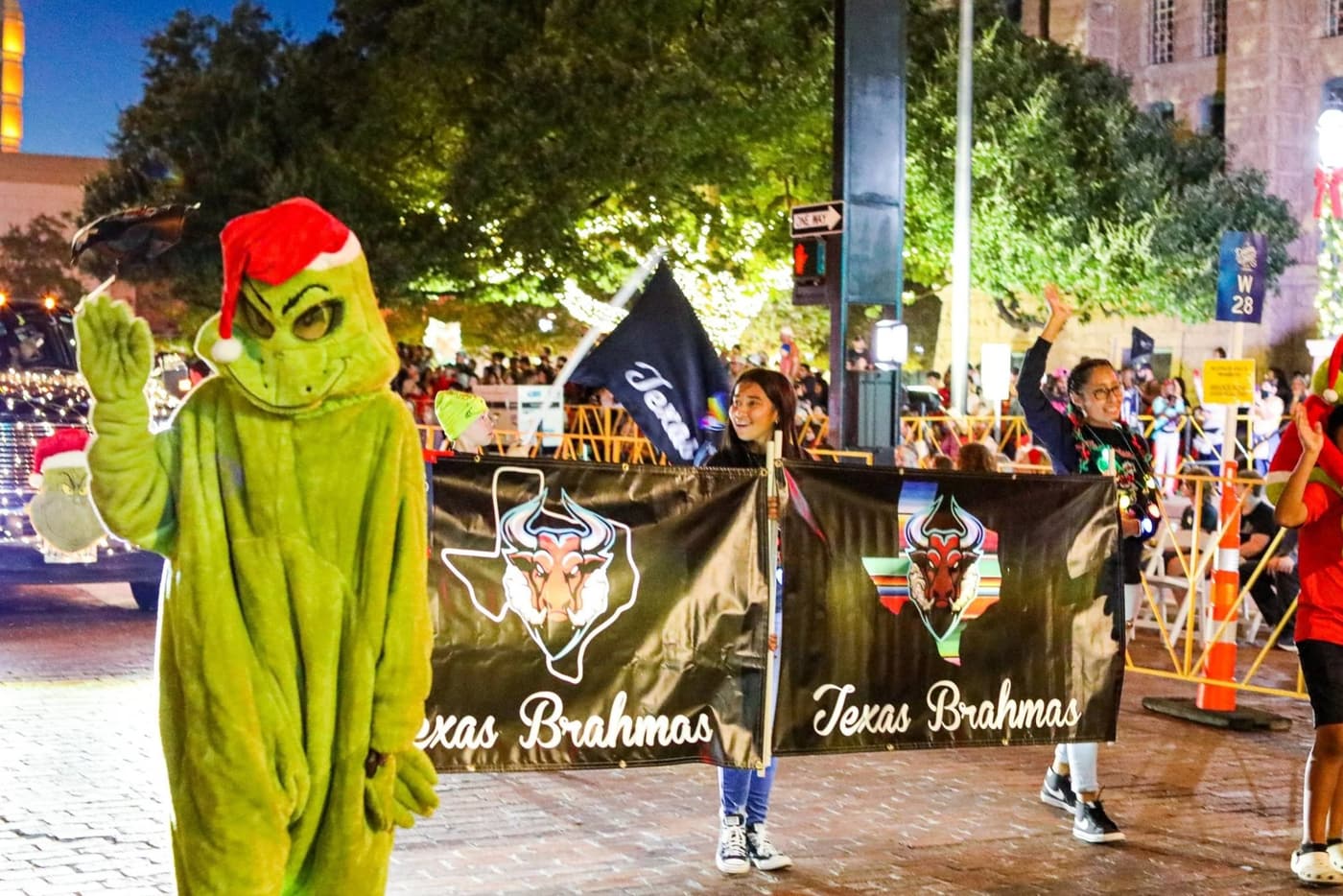 A person in a green Grinch costume wearing a red Santa hat waving, with a crowd of people and festive holiday banners in the background.